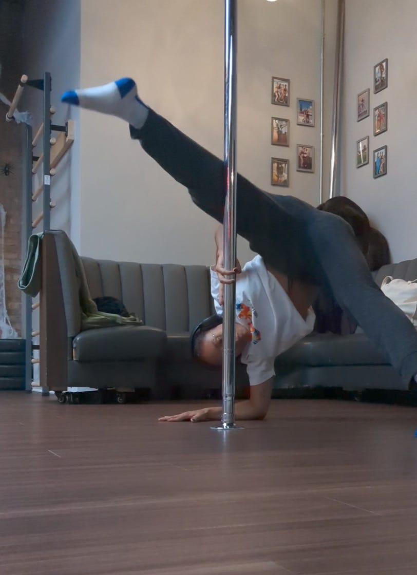 Nathalie is using her right forearm to lift her body up into a half split against a metal pole. She is wearing a white t-shirt. dark gray sweatpants and blue and white socks.