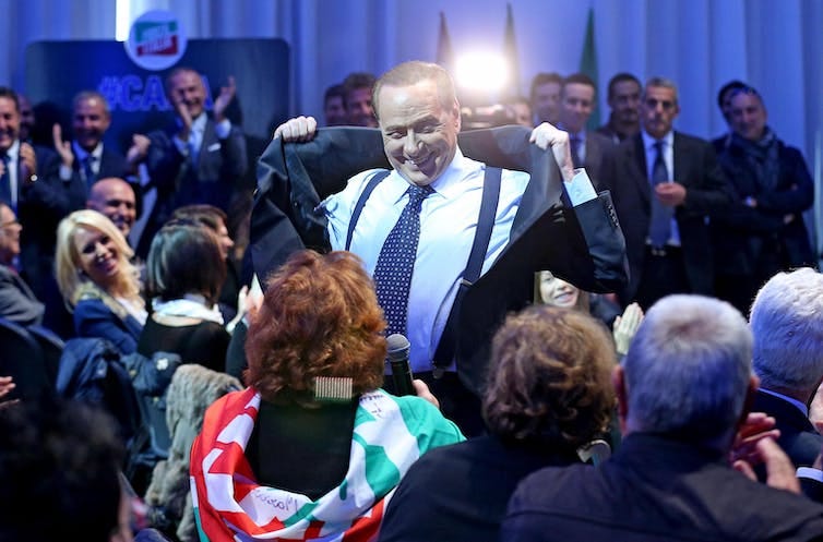 Silvio Berlusconi grinning as he removes his jacket in front of a crowd of people.
