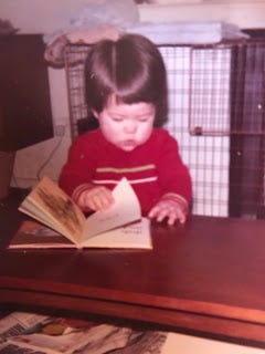 Me, as a toddler. I've got a brown bowl head haircut, and am about 2 years old. I've pulled myself up to the coffee table, and am reading a picture book. I'm wearing a red jumper dress and have a serious expression on my face. The carpet has a brown floral pattern and the walls are beige. It's screams 70s decor.