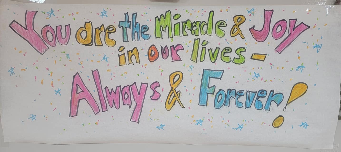 This image shows the hand-painted poster that the author created for her aunt, reading "You are the miracle and joy in our lives, always and forever." The lettering is in pastels, and stars and confetti surround the words.