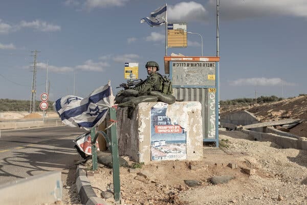An armed soldier standing behind a concrete barrier and sandbags. He is flanked by Israeli flags.