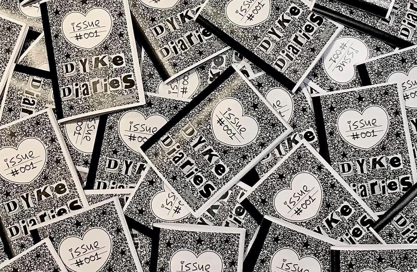 A spread-out pile of the same black-and-white zine issue fills the image. The zine cover has a white heart inside which reads 'issue #001'. Underneath is the title in cut-out letters: Dyke Diaries.