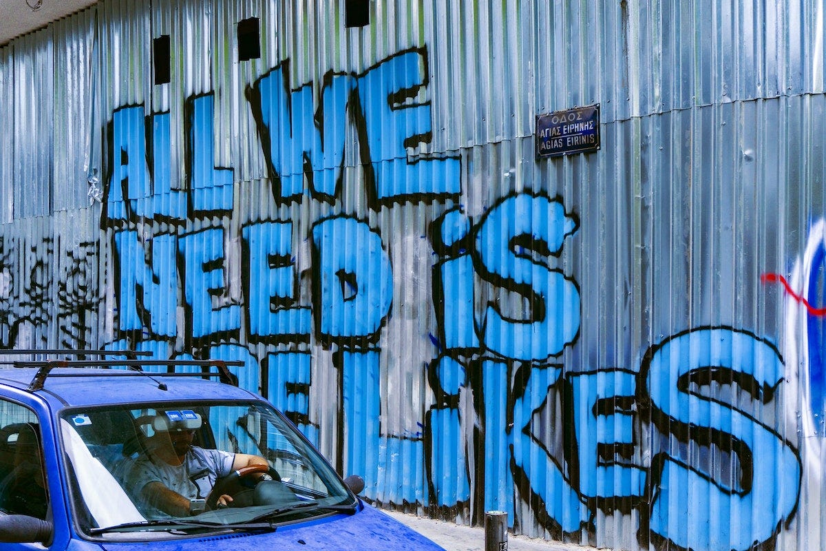 A metal wall painted in big blue letters with the message: All we need is more likes.