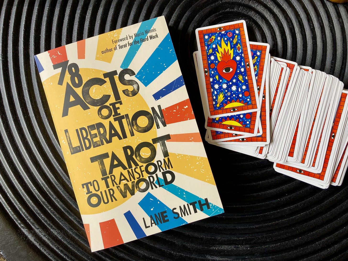 Lane's book, 78 Acts of Liberation, sits next to a tarot deck that spills out over the black table. The book cover shows a brightly colored sunburst with bold black text. The tarot card backs show a flaming heart with an open eye on a royal blue, starry background.