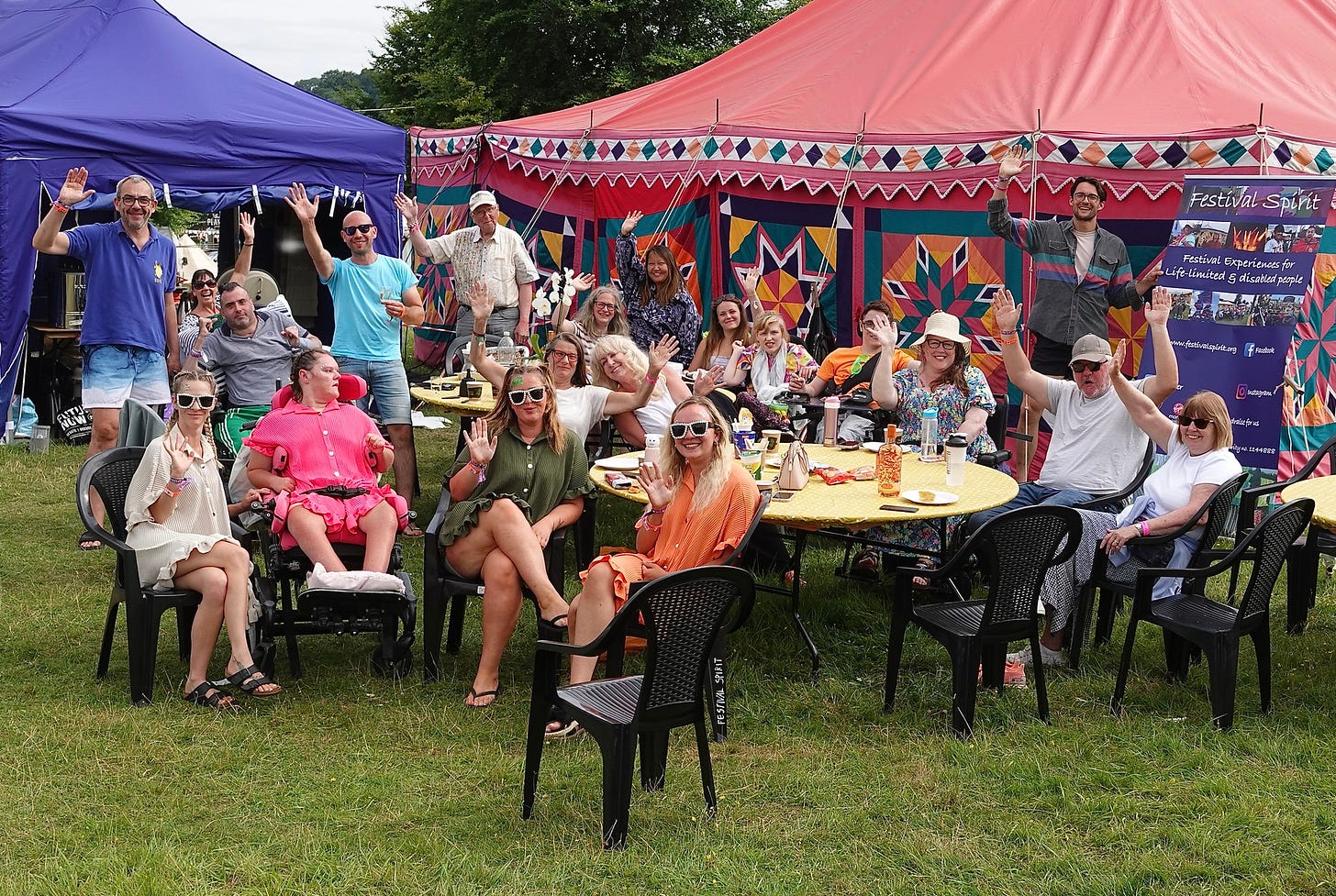 A group of people, some in wheelchairs and others sat.  There are a few people standing.  Everyone is waving at the camera.  In the background is a red brightly patterned marquee and a blue gazebo. Everyone looks happy.