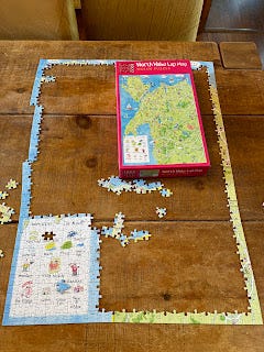A brown wooden table, with the edges of a jigsaw on it. The sides all meet up, although there's a break in the left hand side. In the centre, where there are a few pieces of jigsaw scattered around, the lid of the box is placed. It shows a map of North Wales with illustrated castles, and landmarks.