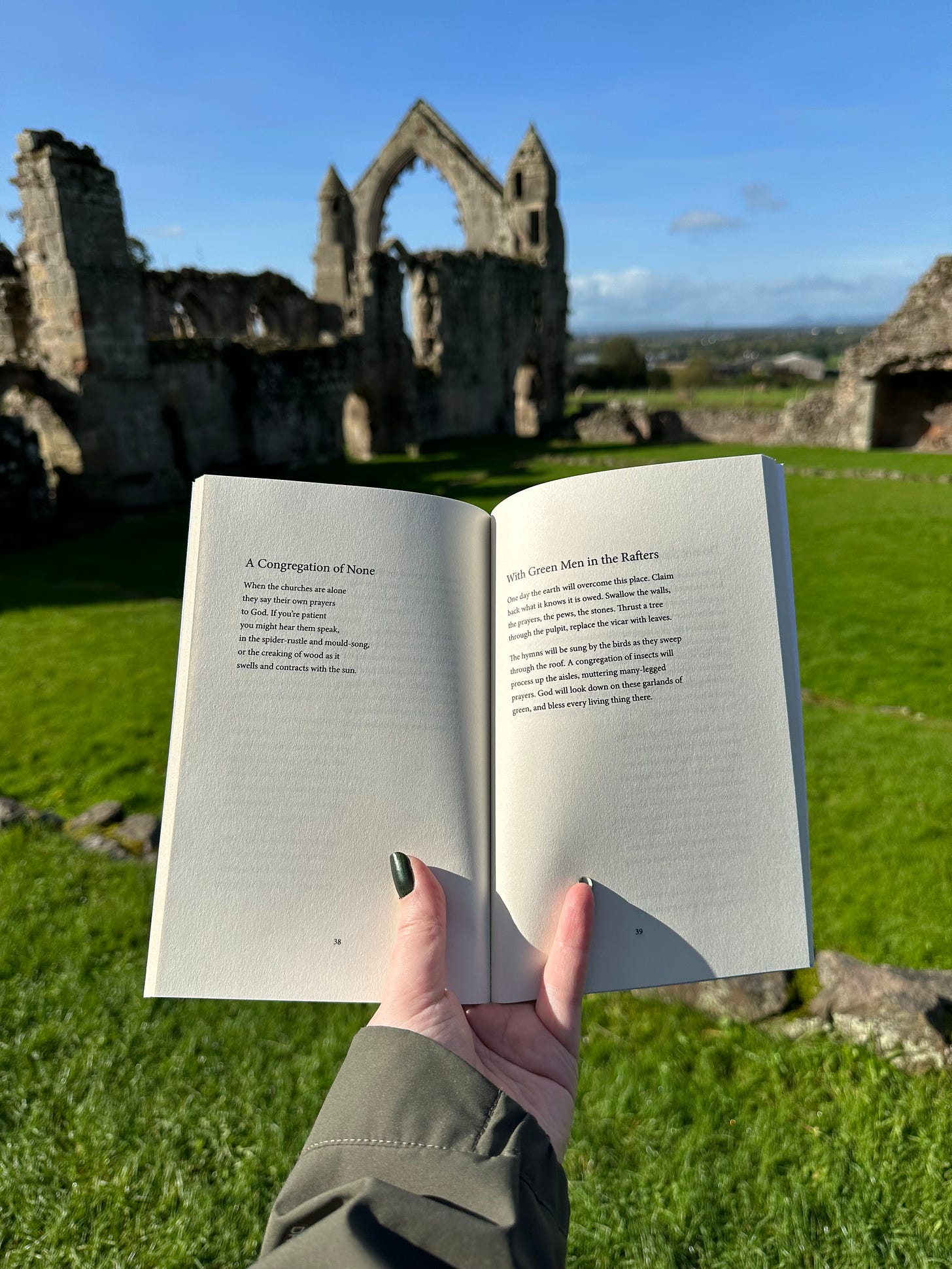 Jay’s book, open to the pages with the two poems I read out, held above the grass and the remnants of stone walls, with the same massive ruined window in the background. The sky is bright blue and the sun is strong.