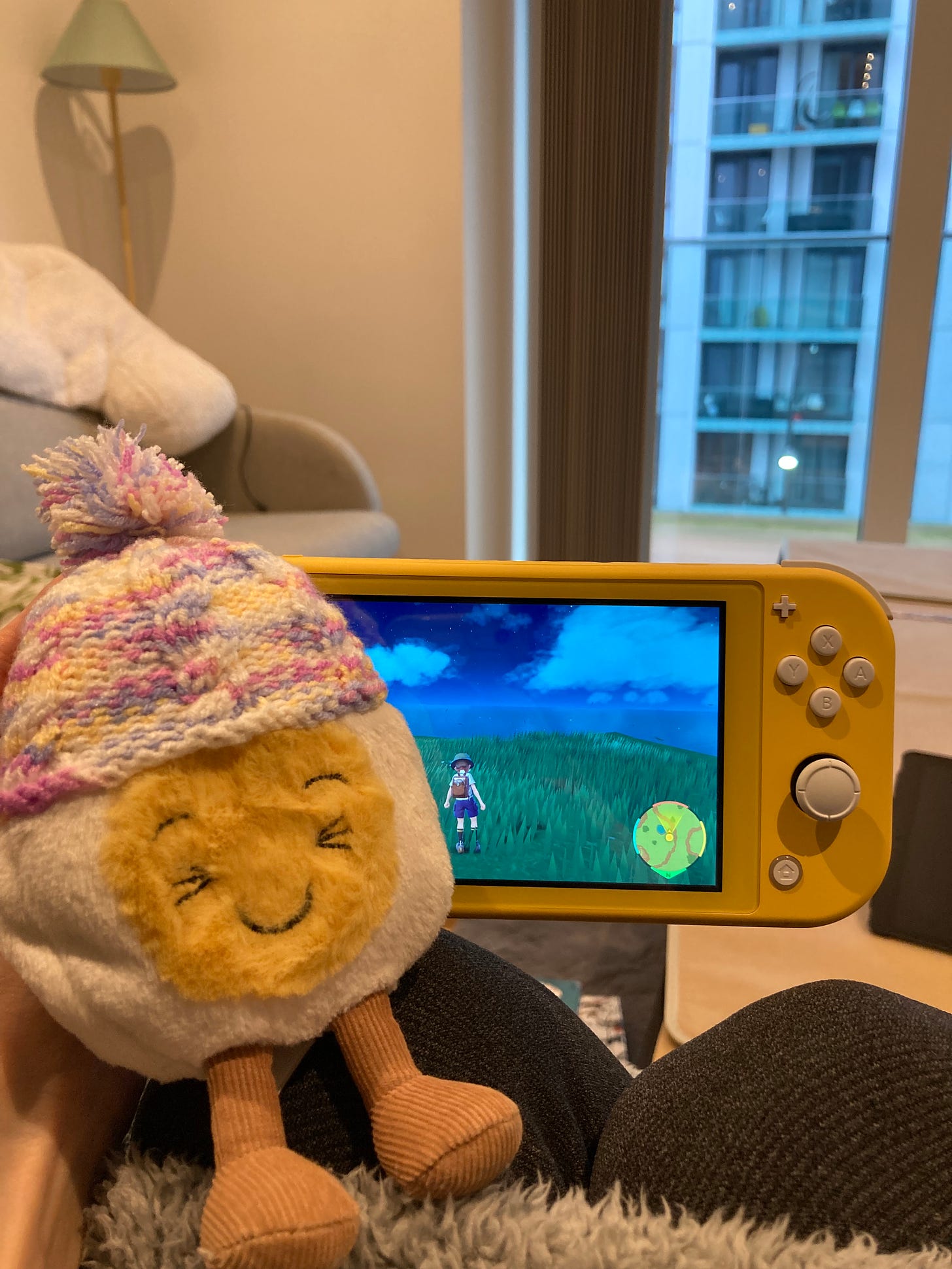 Photograph of dippy the soft toy boiled egg wearing a mini knitted hat, guarding a yellow Nintendo Switch that Beck's hand is holding. The switch displays a pokemon game with the avatar standing on a grassy mountain looking out onto a deep blue sky populated with soft clouds. 