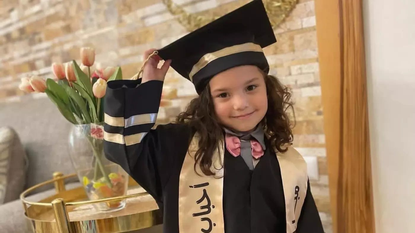 A little girl with dark curly hair and dark eyes stares in the camera with a slight, closed-mouth smile, her head tilted slightly to her right. She is wearing what appears to be a black graduation gown with pale gold satin stripes at the end of the sleeves, a pale gold satin stole with arabic writing on it, and a black mortar board with a pale gold satin stripe and tassel. She holds the sitde of the mortar board with her right hand. In the background you can view what appears to be a living room with a beige/gray couch, exposed brick wall, gold side table, and a vase with tulips