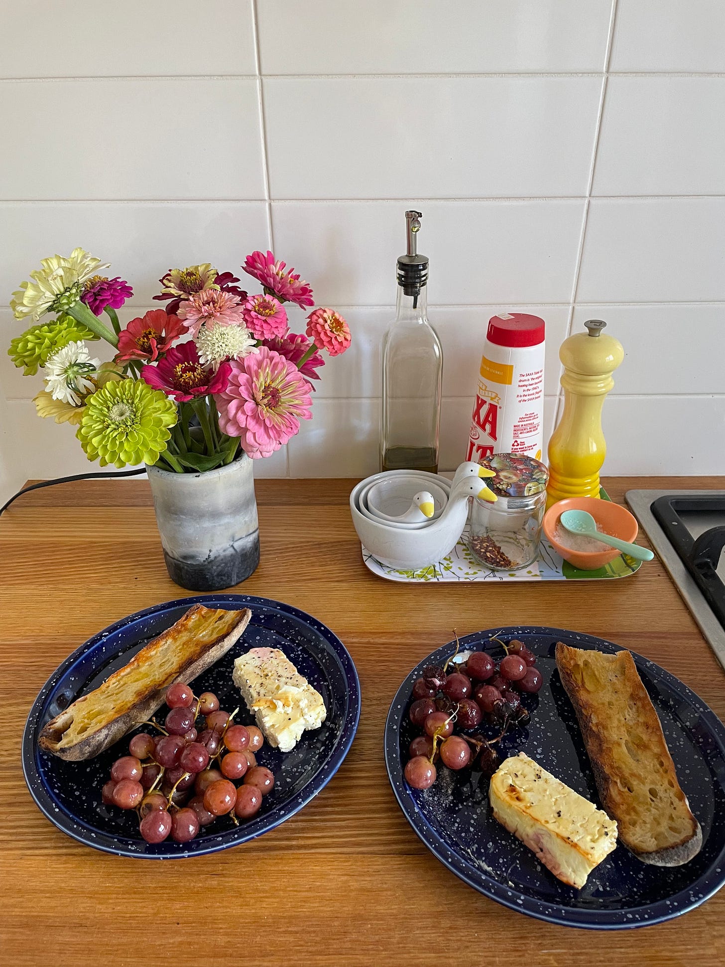 Two plates with roasted red grapes and feta cheese hunks, sitting beside buttered baguettes. It's taken in the kitchen with a small vase of colourful zinnia flowers, and cooking staples like olive oil, sea salt, pepper and chilli flakes in the background.