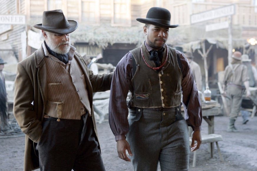 This image shows George Hearst (played by Gerald McRaney) on the left and Odell Marchbanks (played by Omar Gooding) on the right. Both men walk down Deadwood's main thoroughfare.