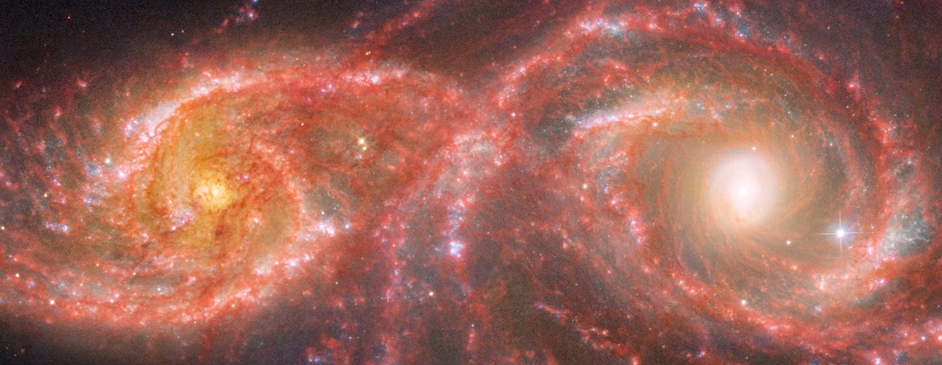 Two brightly coloured galaxies in shades of red, orange, and white, are shown swirling into one another against the deep black of space. 