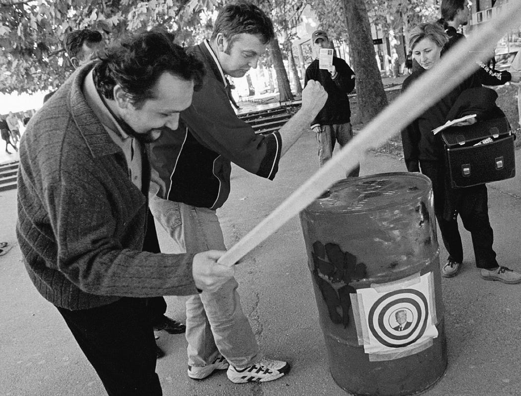 ID: Black and white photo of an Optor street outreach tactic--a passerby pays a dinar to hit a barrel with the president's face on it.