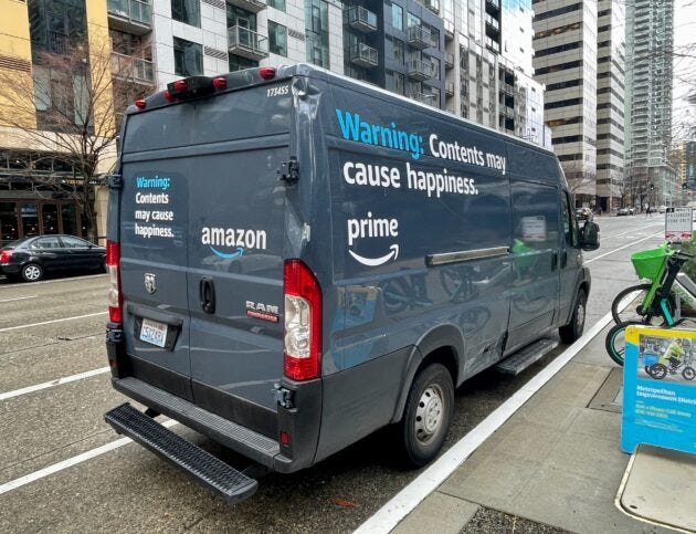 Image shows a blue Amazon delivery van with the words "Warning: Contents may cause happiness" printed on the side and back.