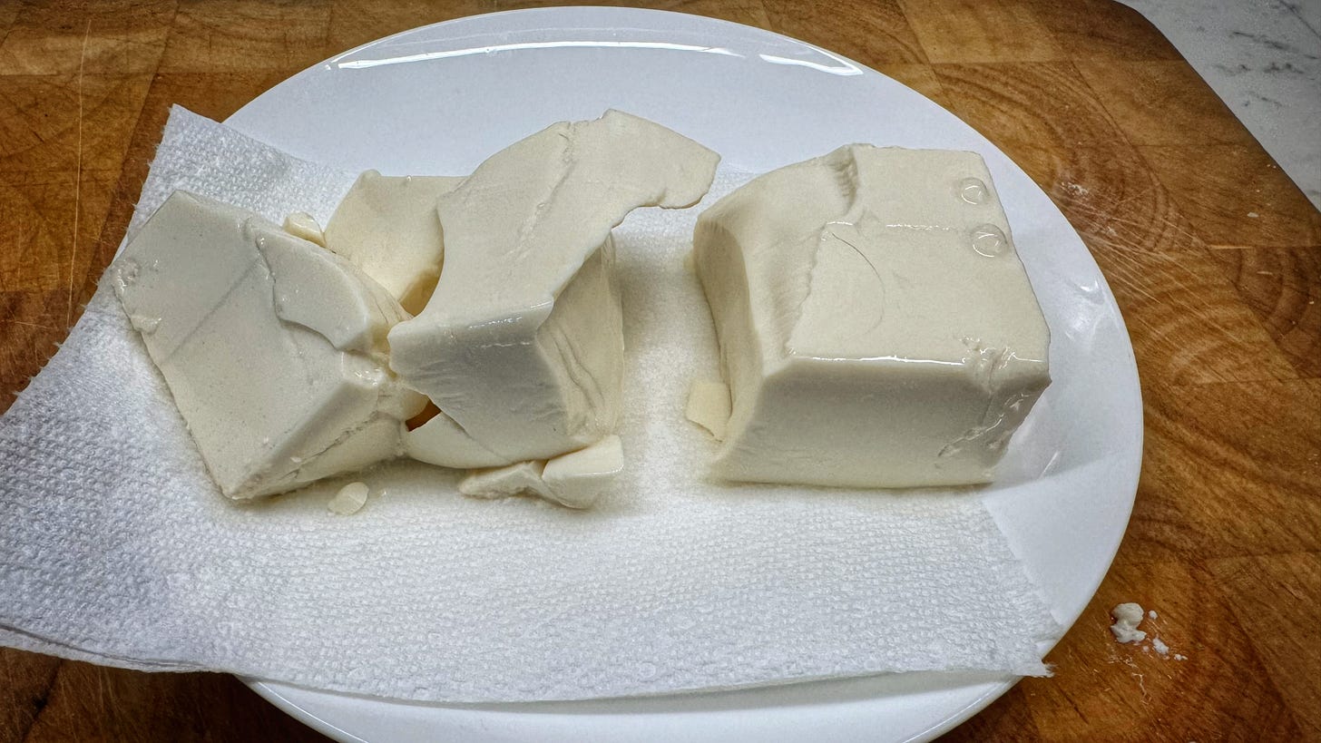 A block of silken tofu draining on paper towel on a plate. The tofu has fallen apart into several large pieces.