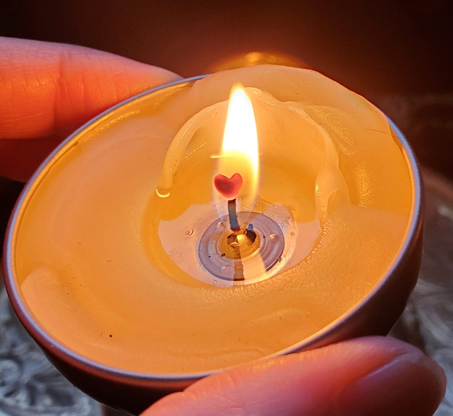 A close up of my hand holding a tiny, burning candle. In the center of the flame, the candle wick has formed the very definite shape of a heart.