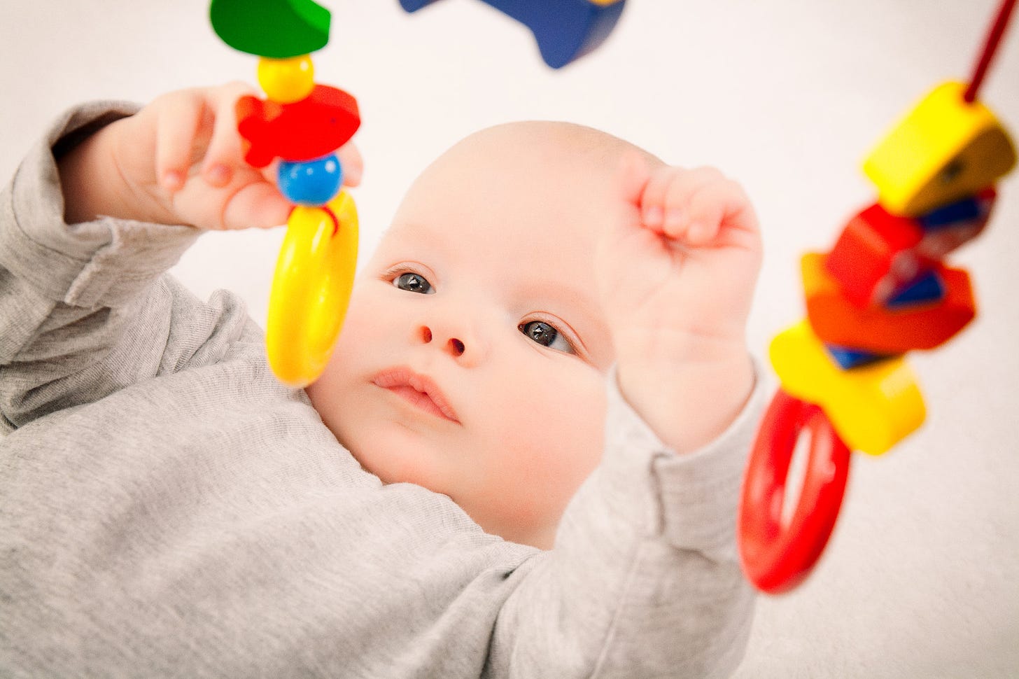 A baby laying on their back on a white blanket and reaching up to touch hanging pieces of a mobile with intense focus.