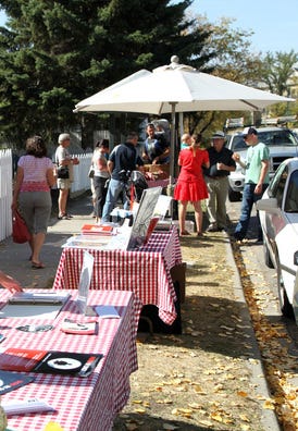 Photo of tables and shade umbrellas set up on a sidewalk boulevard, with people standing around, on a clear autumn day.