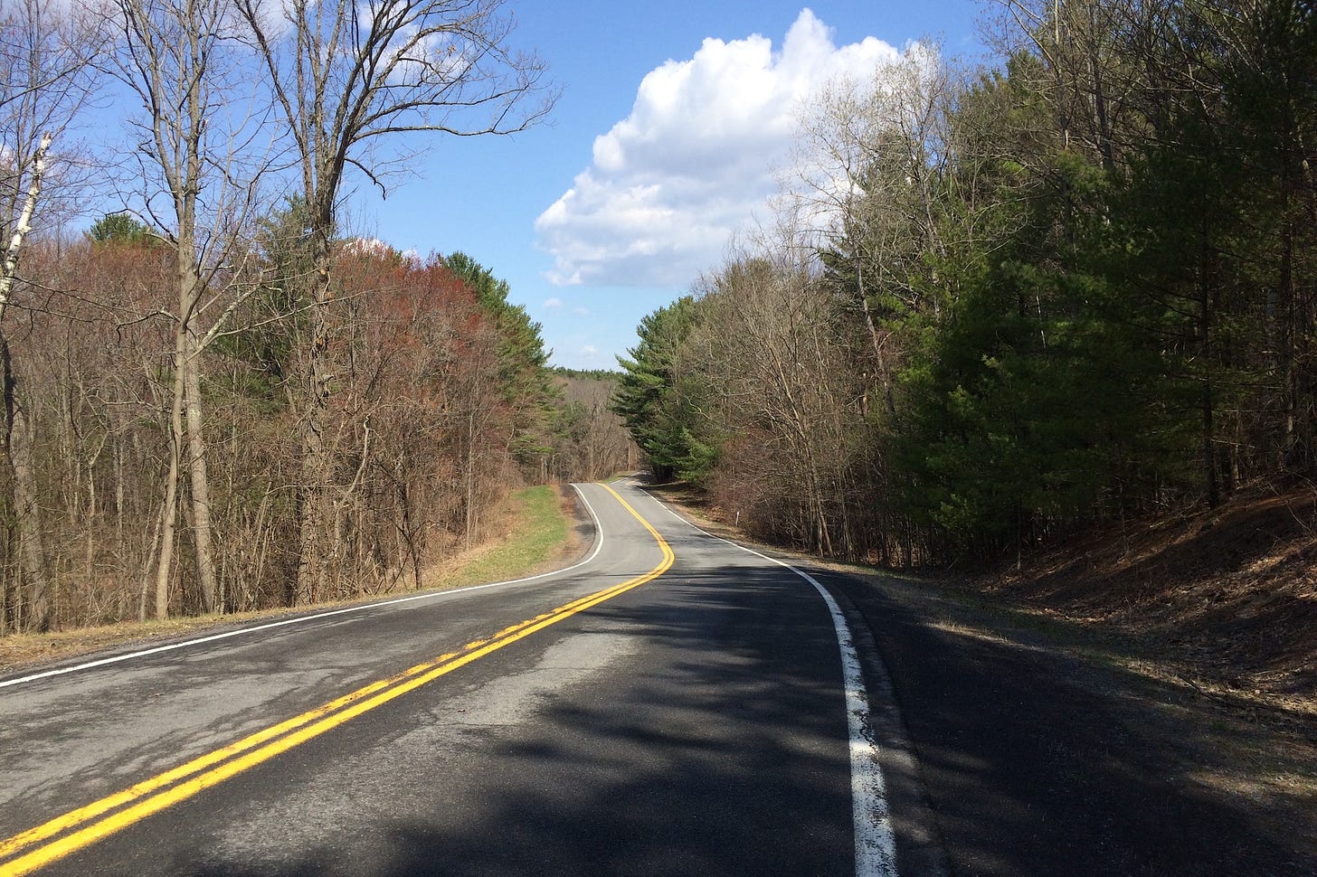 Undulating late fall mountain road, bare trees, but clear blue sky dotted by white clouds. A perfect fall bicyce ride.