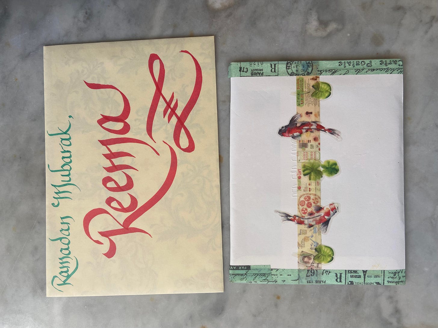 Photo of two decorated envelopes. The one on the left has calligraphy that reads: "Ramadan Mubarak," in green ink and underneath it in larger red caps "Reema." The envelope on the right is sealed with a yellow-patterned washi tape topped with stickers of koi fish and lily leaves and the left and right borders of the envelope are also decorated with patterned pastel blue washi tape. Photo of two decorated envelopes. The one on the left has calligraphy that reads: "Ramadan Mubarak," in green ink and underneath it in larger red caps "Reema." The envelope on the right is sealed with a yellow-patterned washi tape topped with stickers of koi fish and lily leaves and the left and right borders of the envelope are also decorated with patterned pastel blue washi tape.