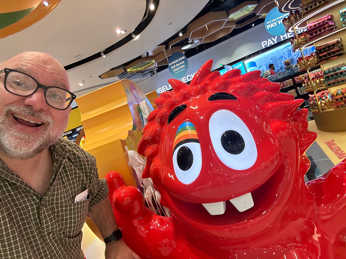 a bald bearded white guy with a goofy grin, next to a 4-foot tall plastic model of a goofy red-colored charactger with two buck teeth, a rainbow eyelid, and just a wacky demeanor overall.