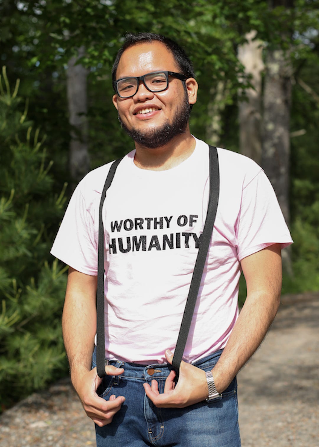 Set Hernandez, a male-presenting person with olive complexion, black hair and beard stands in front of trees, smiling. They are wearing glasses, suspenders, and a pink T-shirt with the text, ‘Worthy of Humanity’.