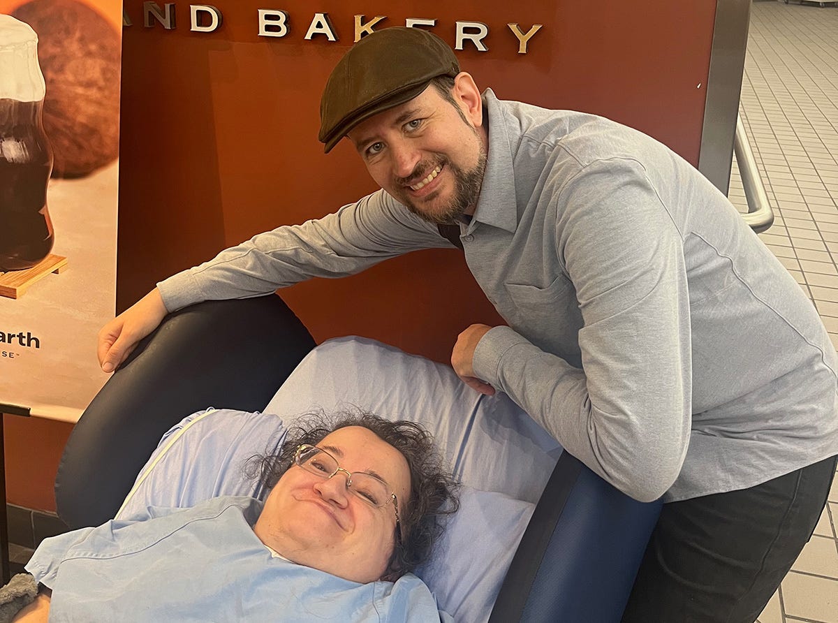 Athena and Stefan at the hospital's coffee shop. Athena is lying in a rolling reclining chair and Stefan is leaning over her. They are both smiling broadly.