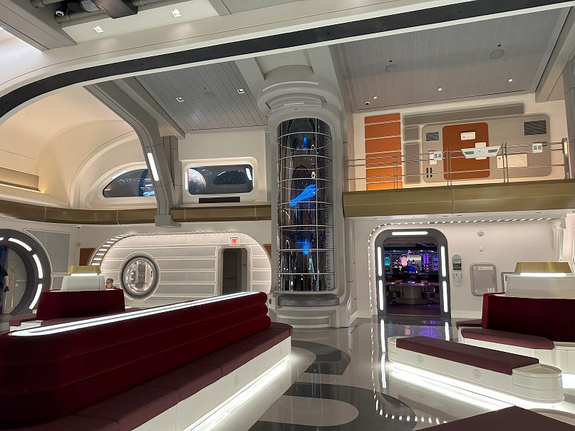 Atrium of the Halcyon Starcruiser: a large open space with a very high ceiling, many viewport windows looking out into space, an arrangement of admittedly shin-bruising benches, and other space-y details