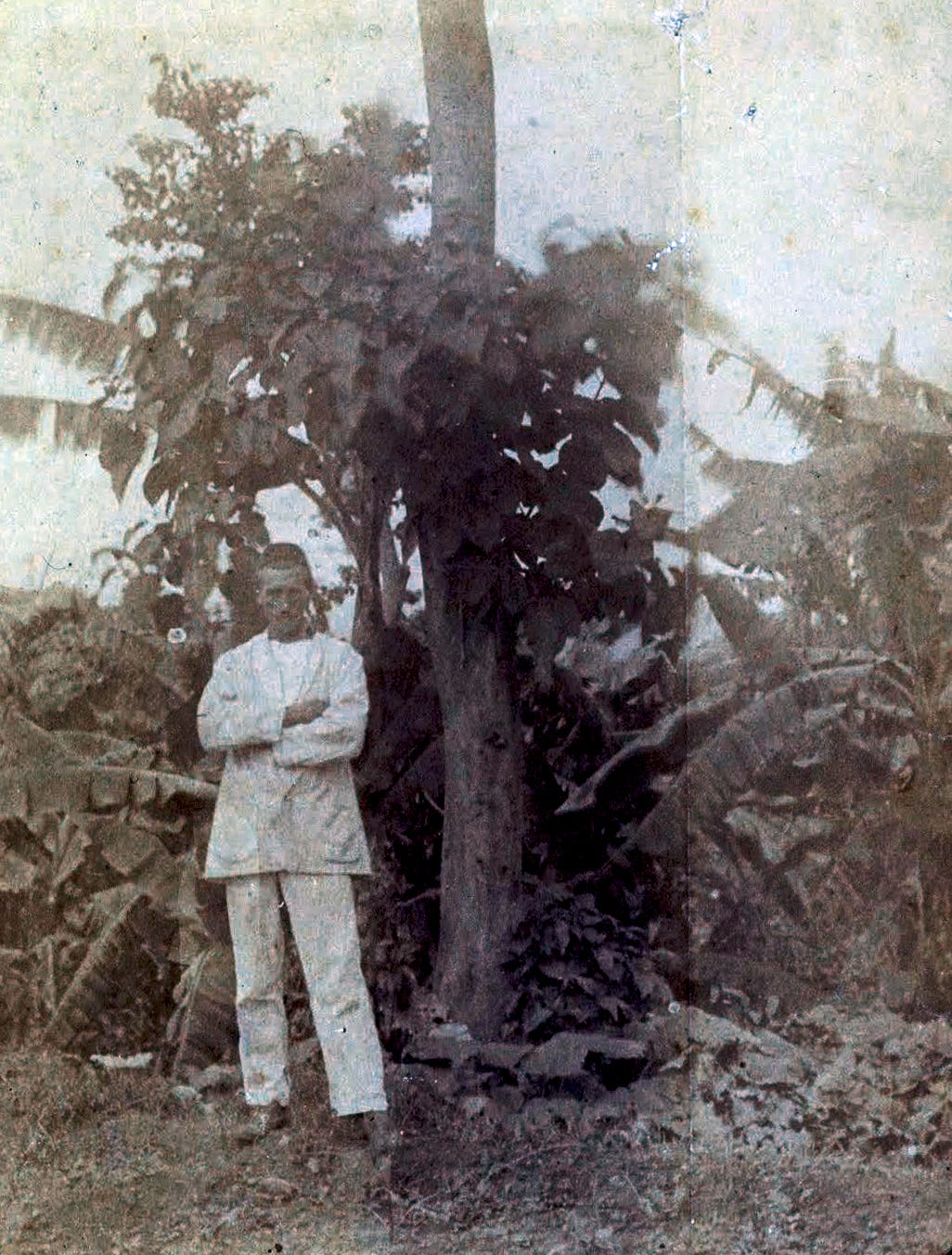 A black and white photograph of a man in white stood in front of a small tree and some various foliage. He is wearing a small fez and has his arms crossed in front of his chest, his legs astride confidently. His face is indistinct.