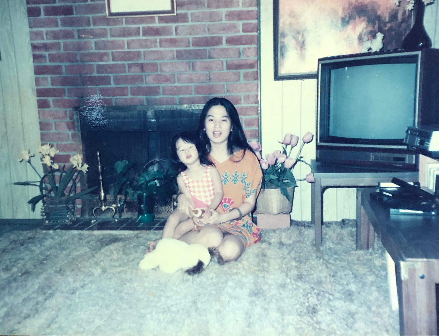 a 1980s photo of a young Asian woman with a little girl in her lap, her daughter. They are in a living room.