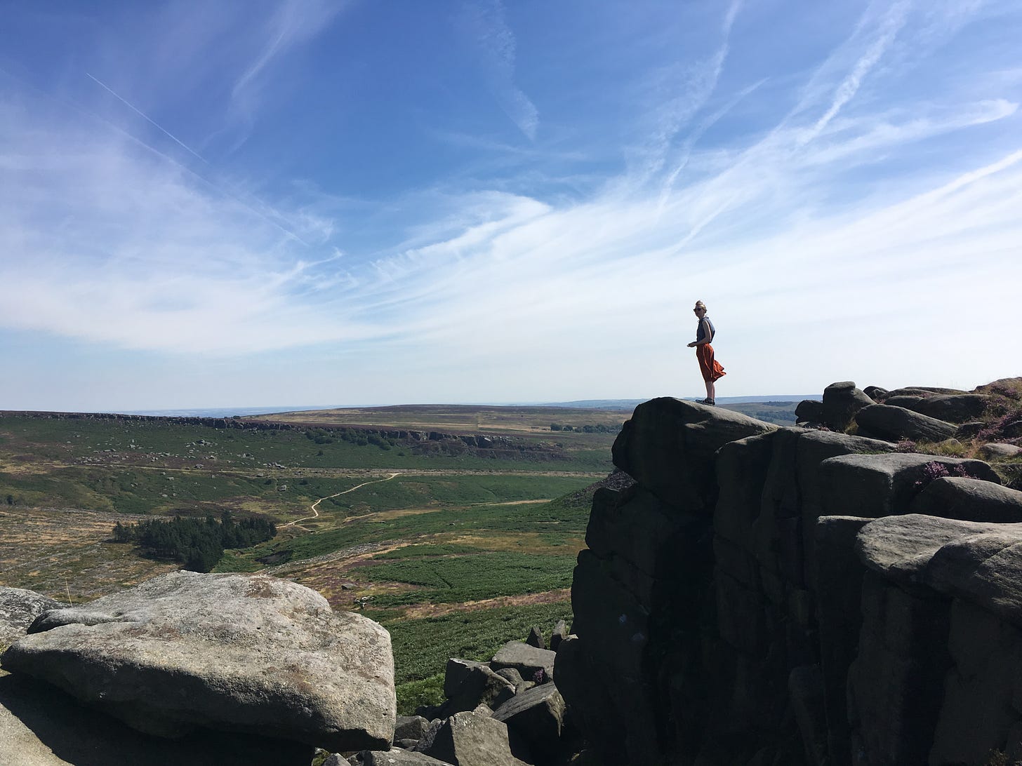 A wide expanse of sky and a flat plain below. in the foreground, large rocks rise up and a young woman in a billowing skirt stands on the edge.