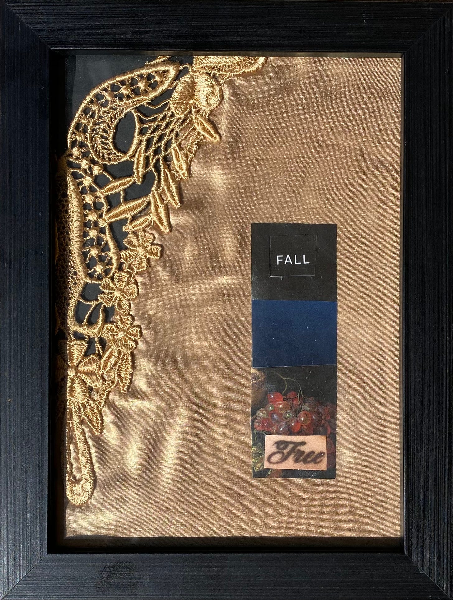 A collage image in the shape of a long rectangle. The top third is black with the word "fall", the middle is dark blue, and the bottom third is an image of a still life painting of grapes with a tattoo of the word "free" on top. The collage is on top of a piece of a gold silk and lace pajama top. Contained in a black matte frame.