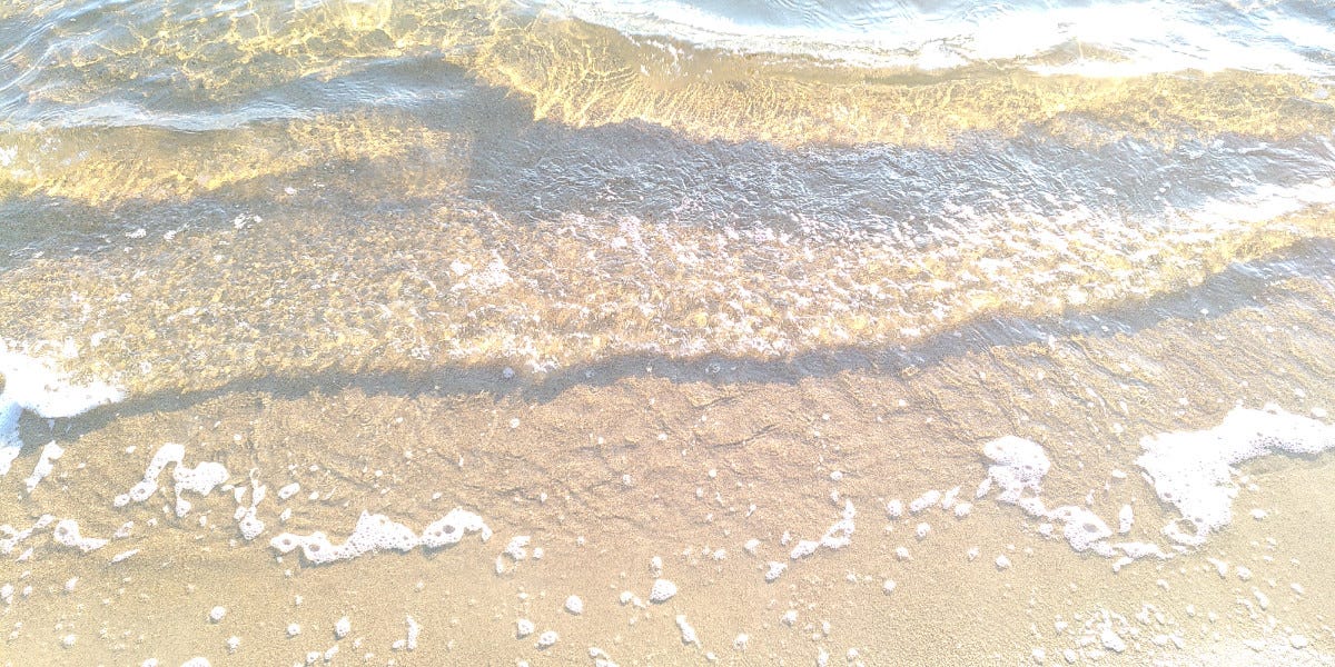 photograph of a sandy beach, with transparent waves crashing against it, white foam forming after the waves, with sunlight lighting up the water