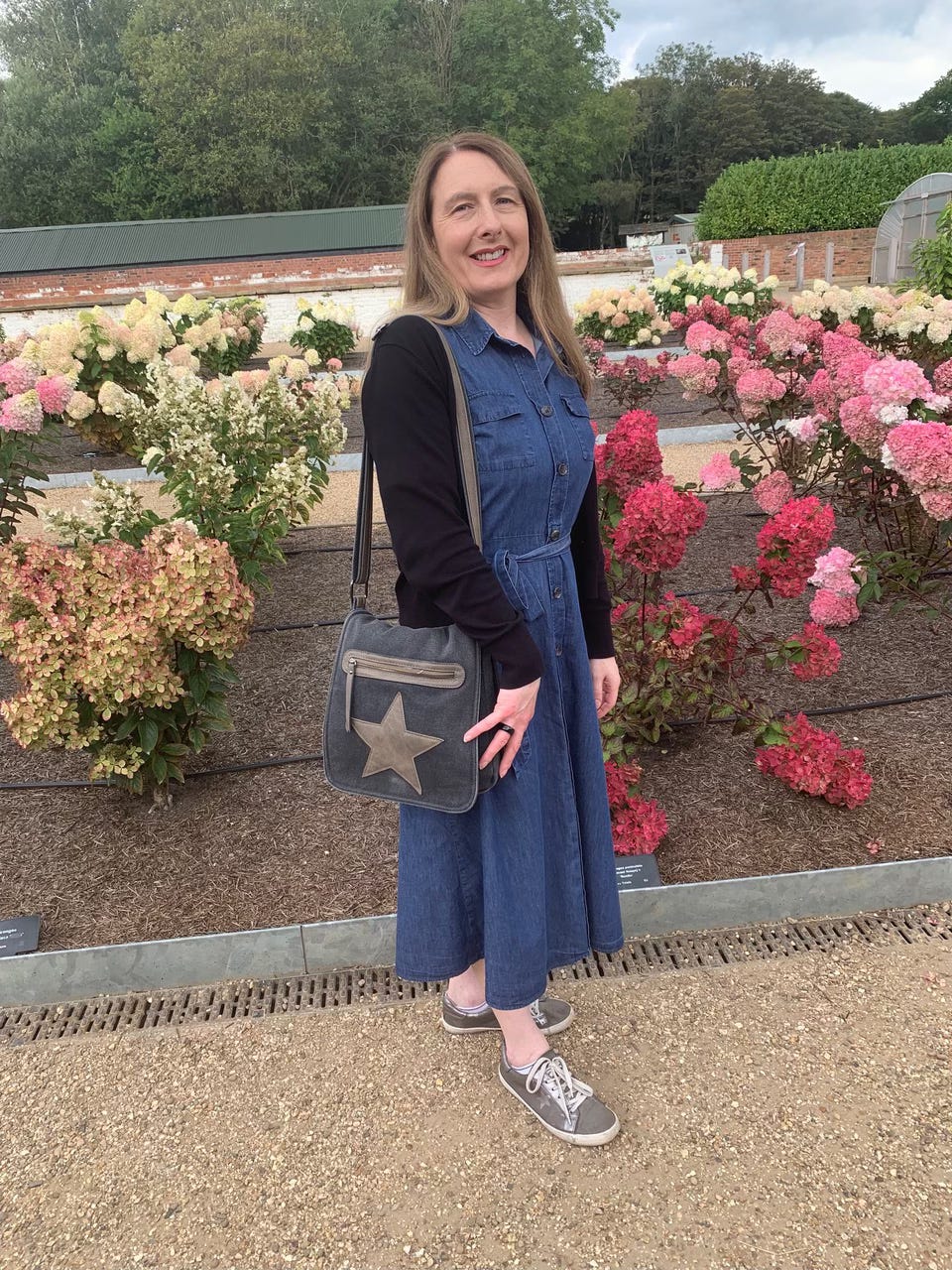 A woman in a denim dress stands in front of a flower garden