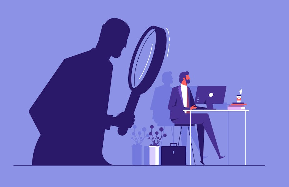 An illustration of someone working at a desk, in a business suit, while a shadowy figure watches, unseen, holding a giant magnifying glass