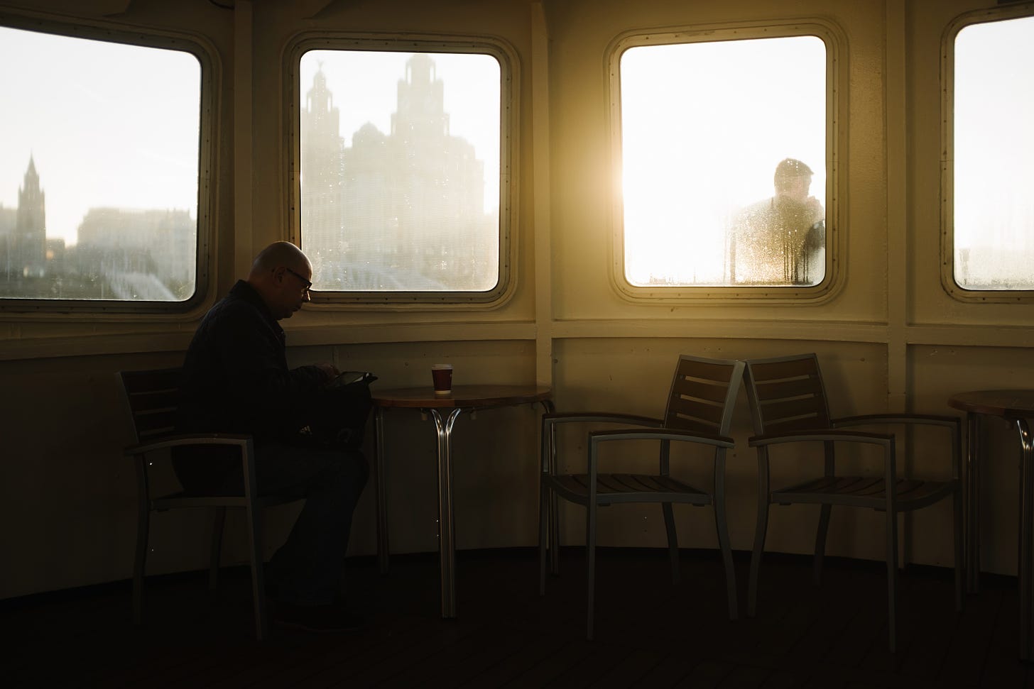A man sits at the front of the ferry with a coffee. It is sunny, early morning, and the light is pouring through the window. The Liver Building is outside.