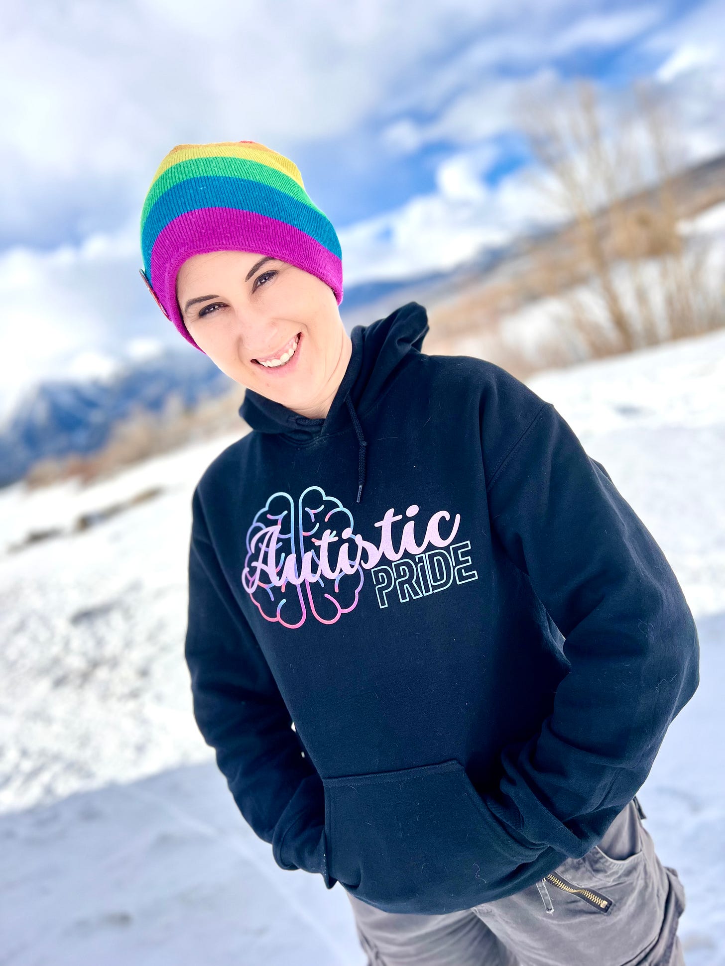 Photo of Lyric on their trip to Colorado. It is winter, and snow is on the ground around them. They wear a rainbow beanie, and black hoodie, with the word Autistic Pride on a brain icon in lavender and teal, with a pop of magenta. Their outfit is pulled together with cargo pants that were not quite in fashion… yet. 