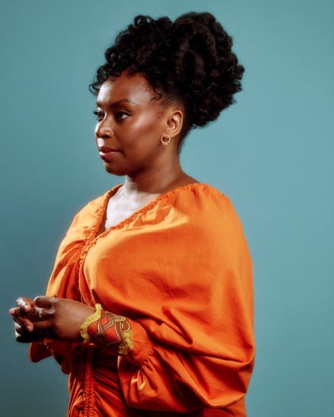 Portrait of Chimamanda Ngozi Adichie with her hair piled on top of her head and wearing a burnt orange top and holding her hands in front of herself