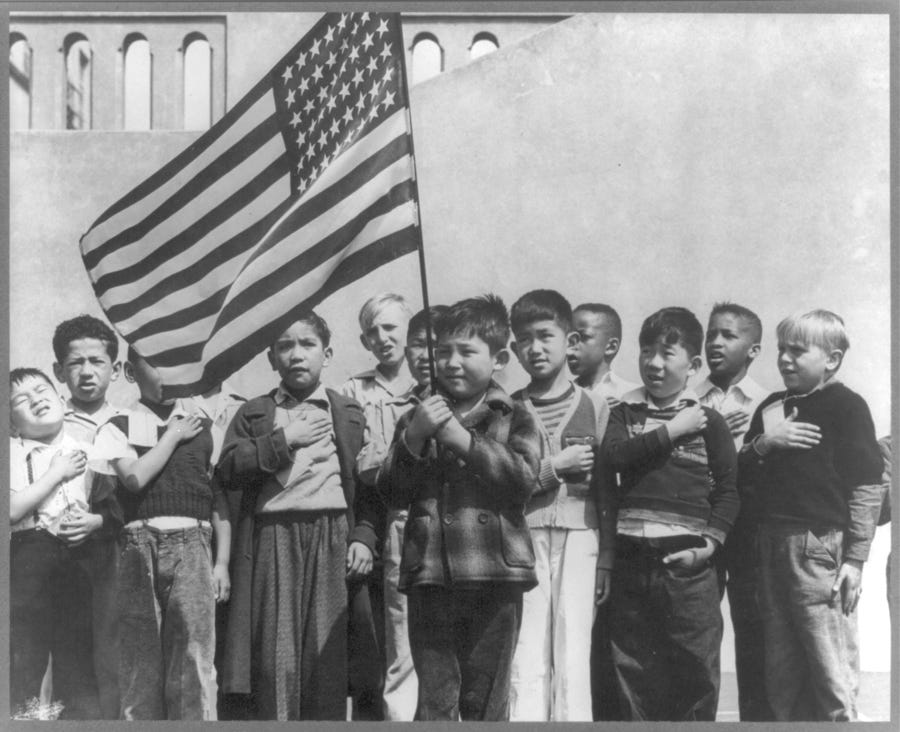 Historic black and white photo of a multiethnic group of elementary schoo kids saluting the flag outside their school in 1942