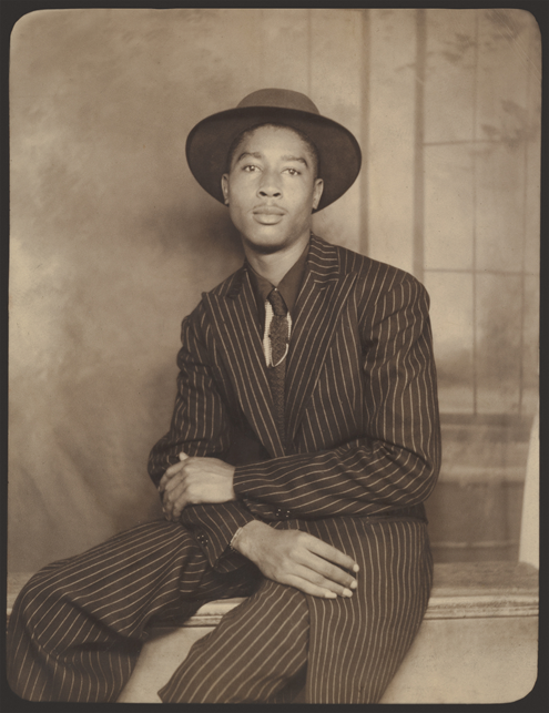 A man in a fine suit and hat sits posed with his arms crossed