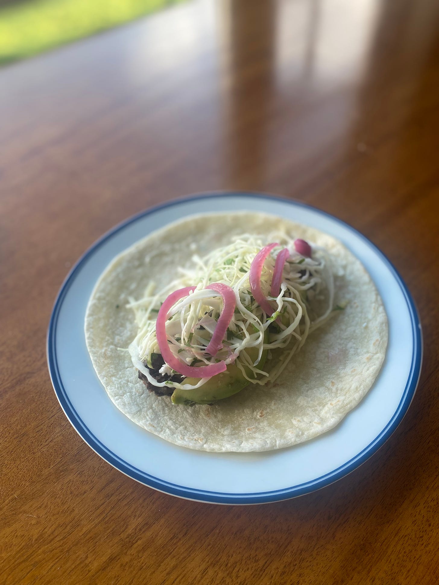 A white flour tortilla with the taco filling described above, black beans and avocado and a sprinkle of feta just visible beneath a cabbage slaw and pickled red onion slices.