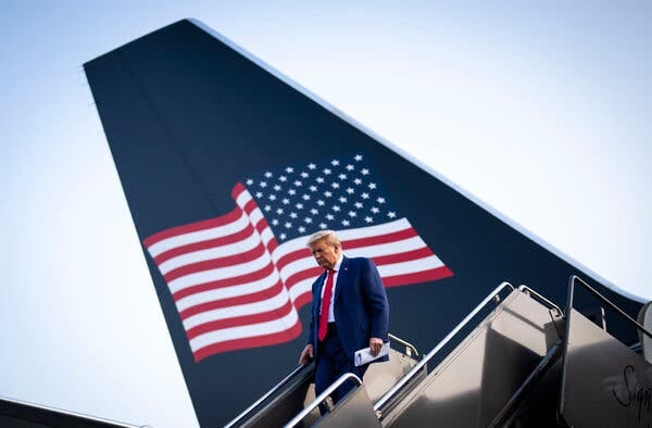 Former President Donald J. Trump, in a suit and red tie, descending the stairs of his plane, with an American flag on the tail behind him.