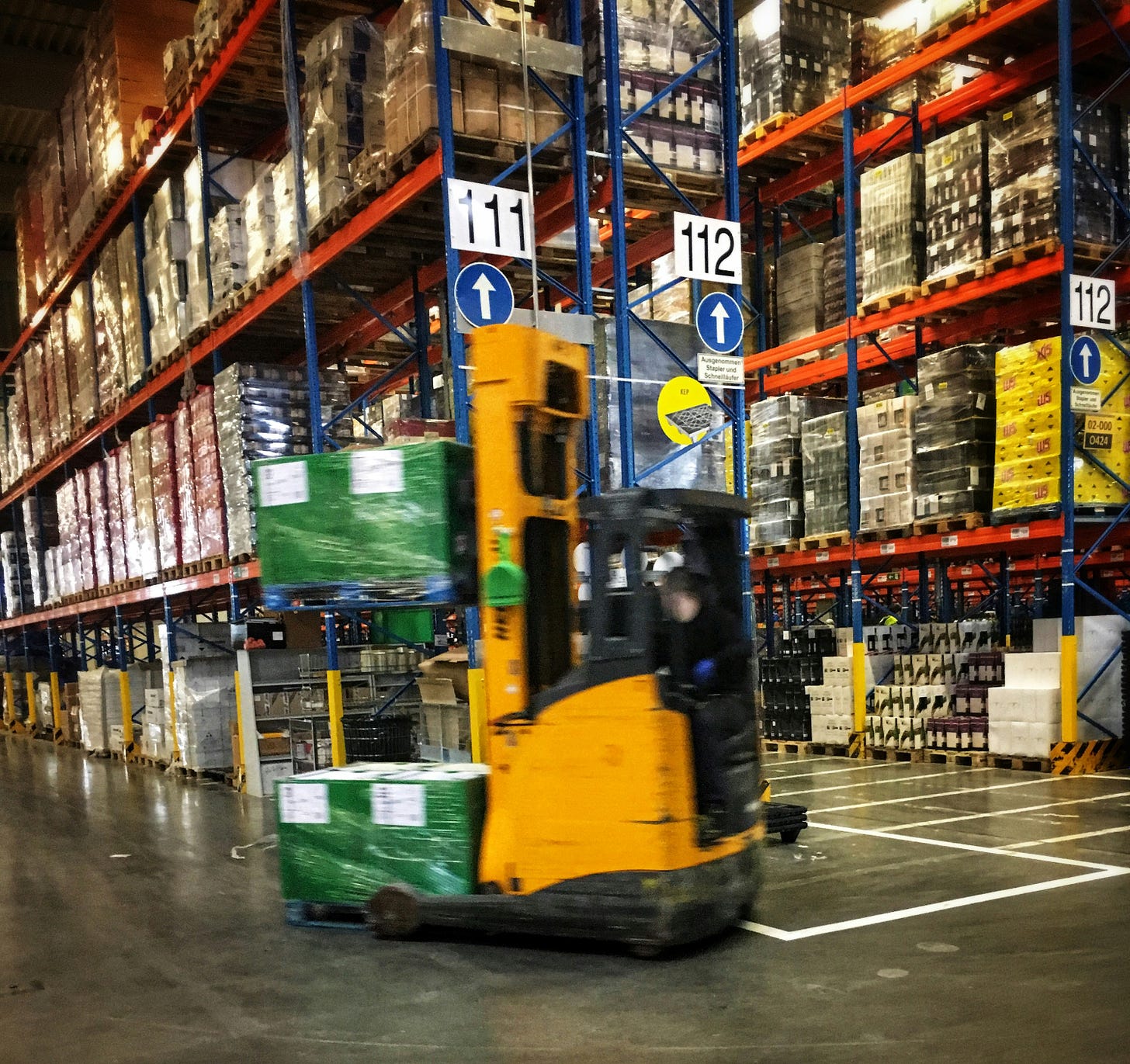 a forklift moves quickly through a large warehouse full of supplies. the forklift is blurry, perhaps due to motion. the shelves in the background are stacked high with pallets.