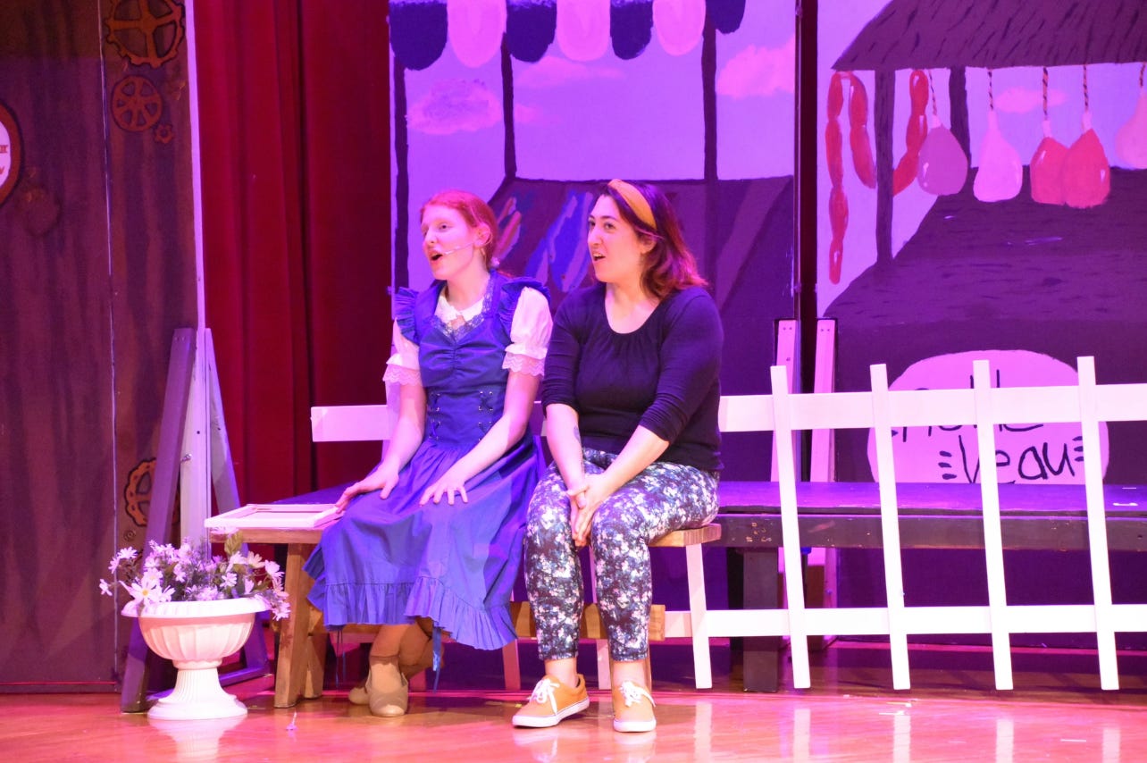 A girl in a blue dress sits next to a woman in a blue sweater and printed pants. Both are seated in front of a fence on a stage, singing. 