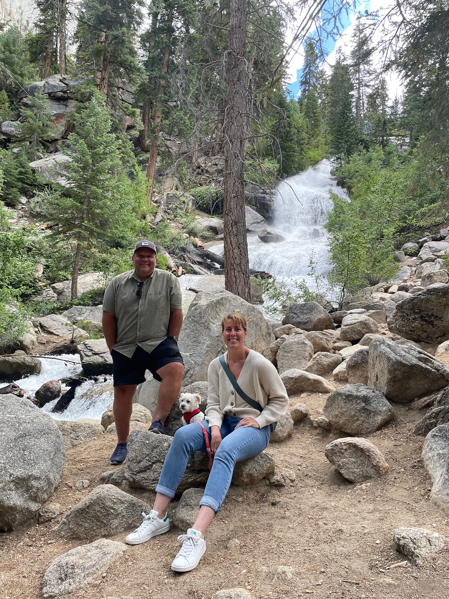 Mickey, Julie, and their dog Ellie at the base of a waterfall, surrounded brgreen trees and boulders in whitney portal