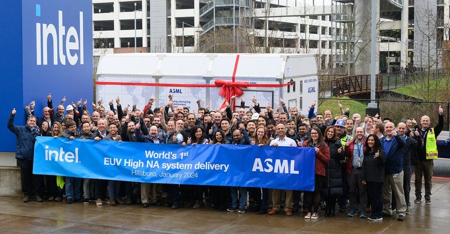 A crowd of Intel and ASML employees hold up a blue banner with Intel and ASML logos that reads "World's 1st EUV High NA system delivery Hillsboro, January 2024." A large white box with a red bow on it sits in the background. 