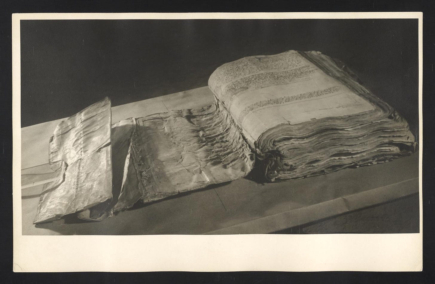 A black and white image of a huge old book with hundreds of mouldering pages opened to the first page.