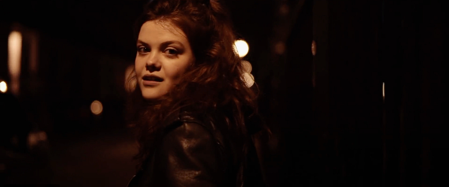 Georgie Henley turns to look directly at the camera. She is bathed in orange-toned light and a few more out-of-focus streetlights appear in the background.