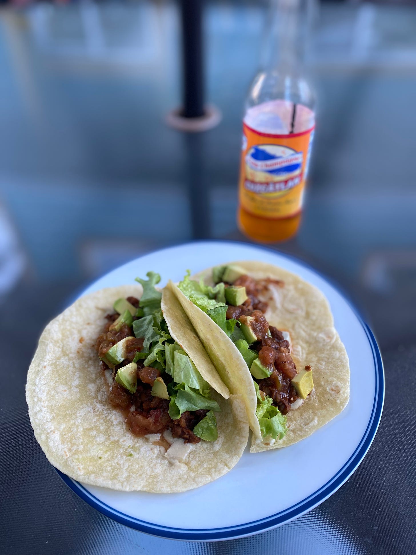 Two tacos in soft shells, each with lentils, salsa, avocado, and lettuce visible. A bottle of Mexican soda sits in the background.