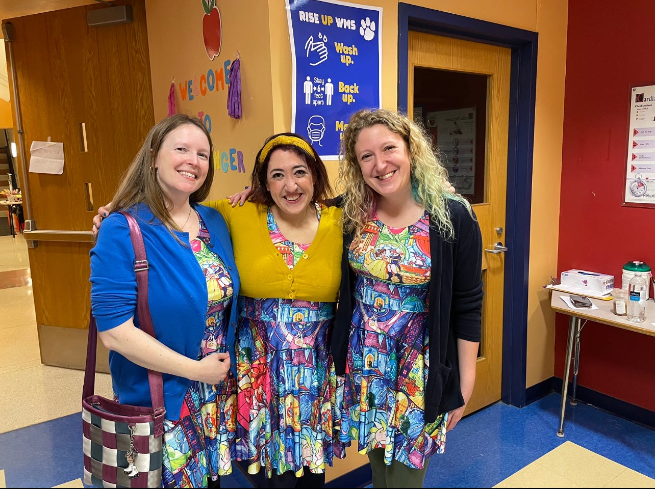 Three women wearing the same Beauty and the Beast dress are standing together with their arms around each other in a school lobby. 